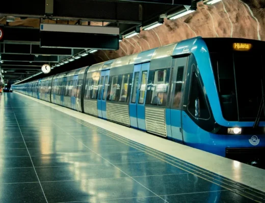 A modern blue and silver metro train stopped at a station with a rock-hewn tunnel ceiling, empty platform, and reflective floor.