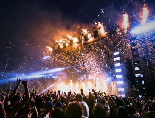 A massive crowd at the Rolling Loud music festival at night, their hands in the air, facing a huge stage dramatically lit with bright lights, fire pyrotechnics, and falling confetti.