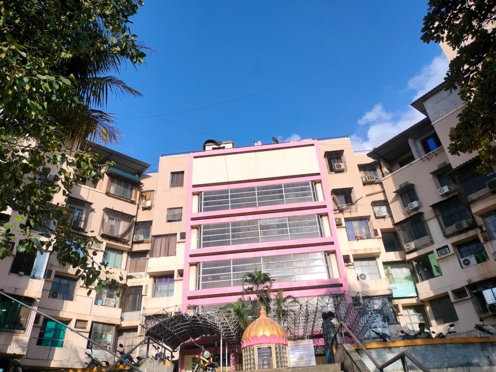 The courtyard of a BIMA complex building, featuring a distinct pink facade and a small golden-domed temple, showing the blend of commerce and culture.